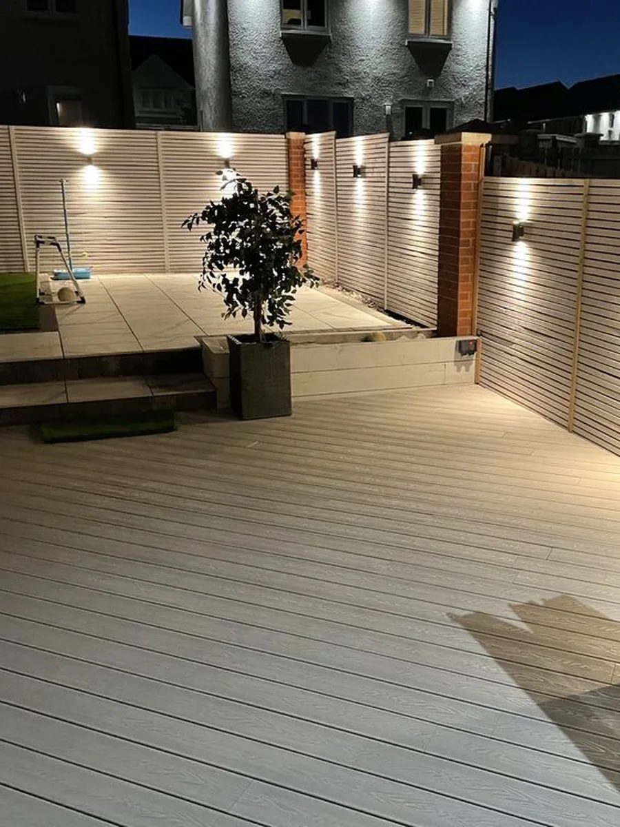 Garden decking at dusk with wall lights on the slatted fence and the planters lit